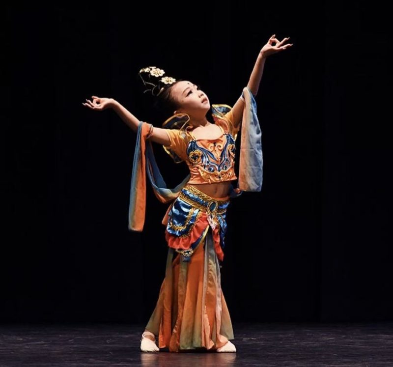 A young dancer in an orange and blue Dunhuang costume with flowing ribbons, arms raised, on a dark stage